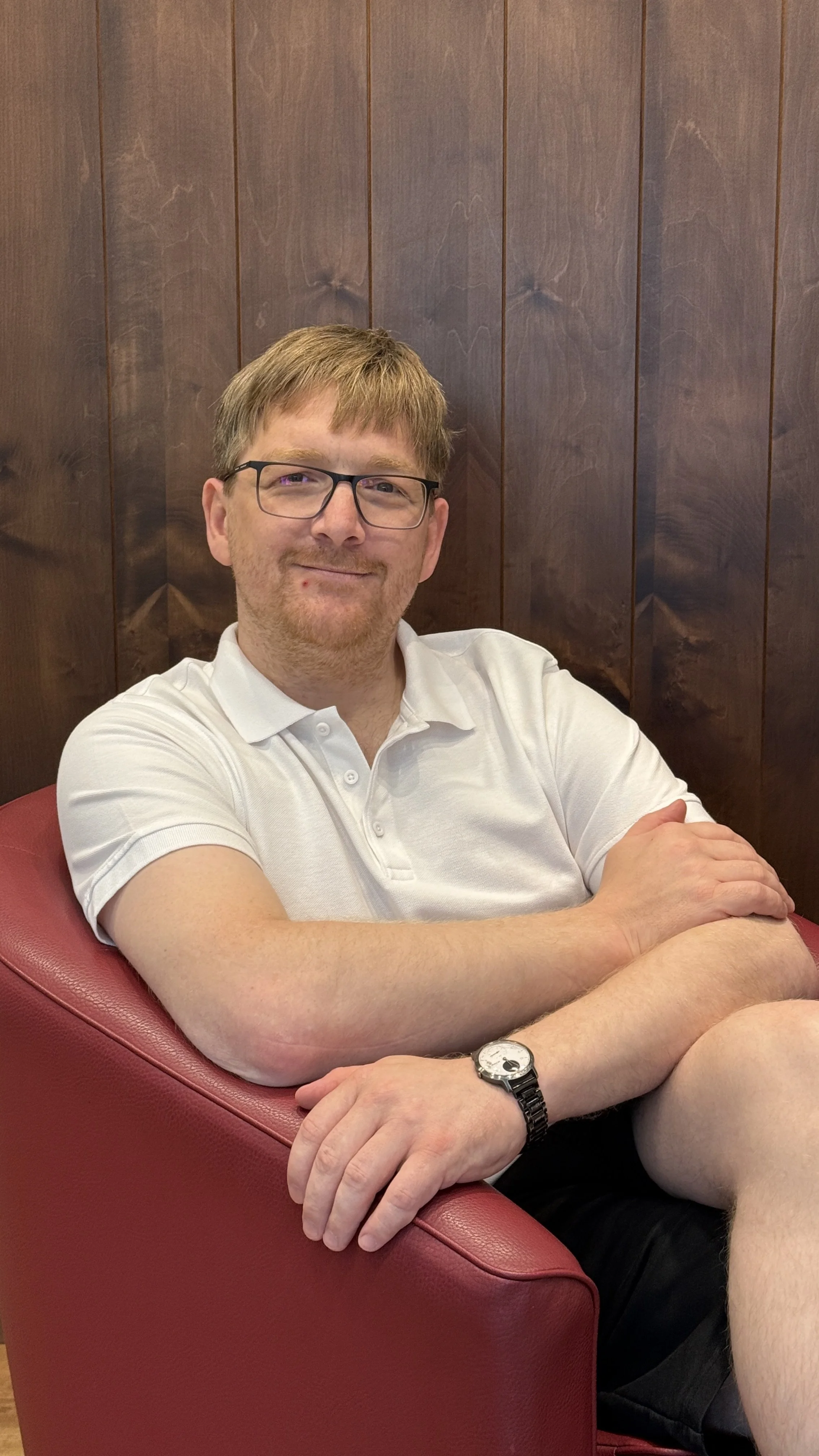 Ein Mann mit Brille in einem weißen Polo-Shirt sitzt entspannt auf einem roten Sessel vor einer Holzvertäfelung.
