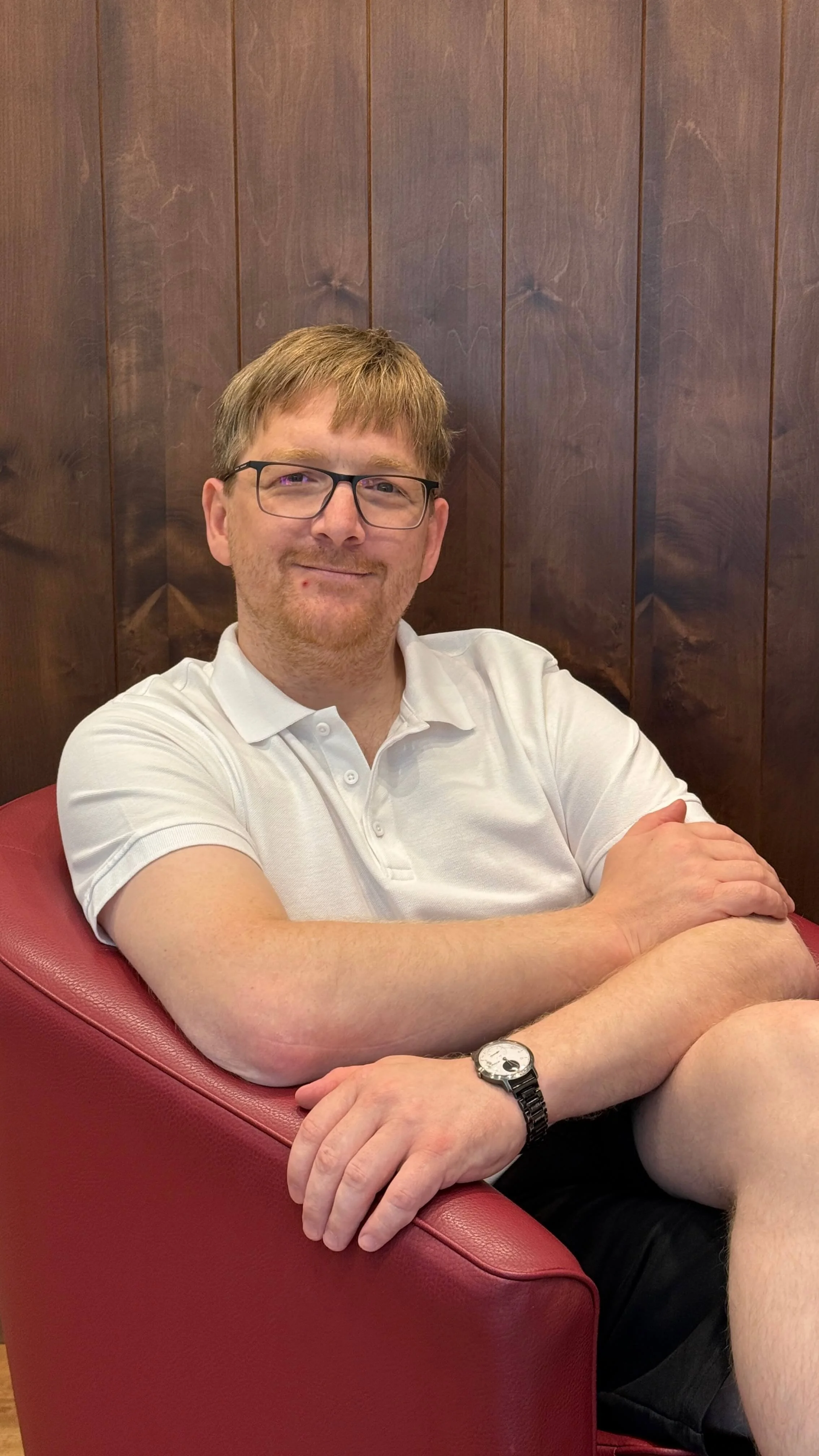 Ein Mann mit Brille, weißem Polo-Shirt, schwarzer Shorts und einer Armbanduhr sitzt auf einem roten Sessel vor einer dunklen Holzvertäfelung, lächelt in die Kamera.