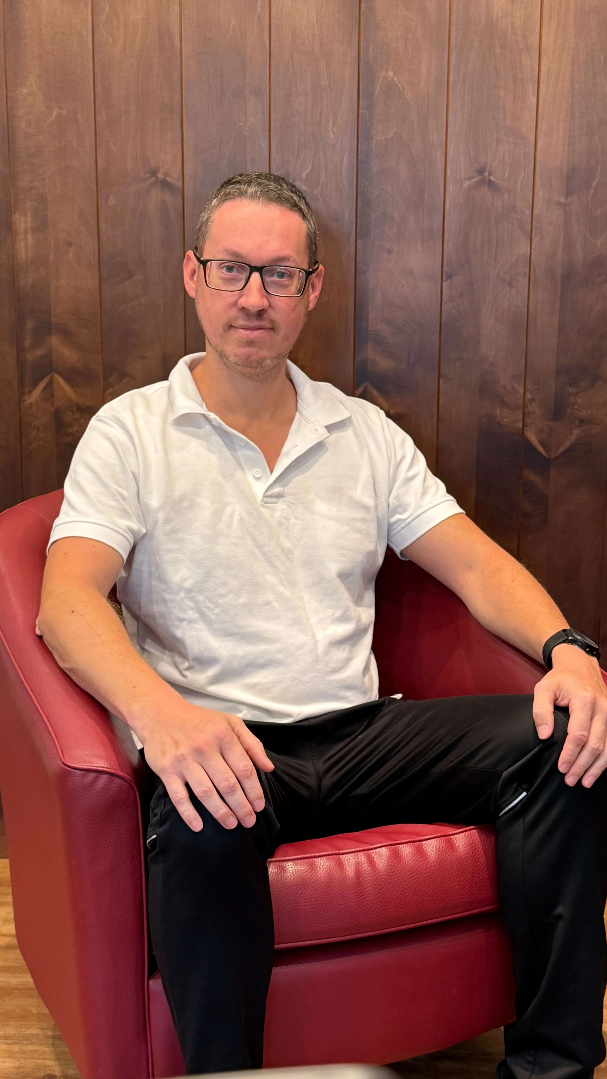 Ein Mann mit Brille in einem weißen Polo-Shirt und schwarzen Hosen sitzt in einem roten Sessel vor einer Holzvertäfelung.