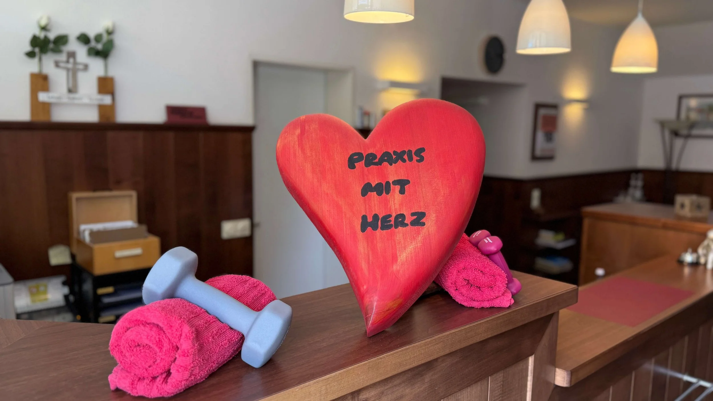Herzförmiges Dekorationsstück mit der Aufschrift 'Praxis mit Herz', eine kleine Hantel und ein Handtuch auf einer Holztheke in einer Praxis oder einem Behandlungsraum.