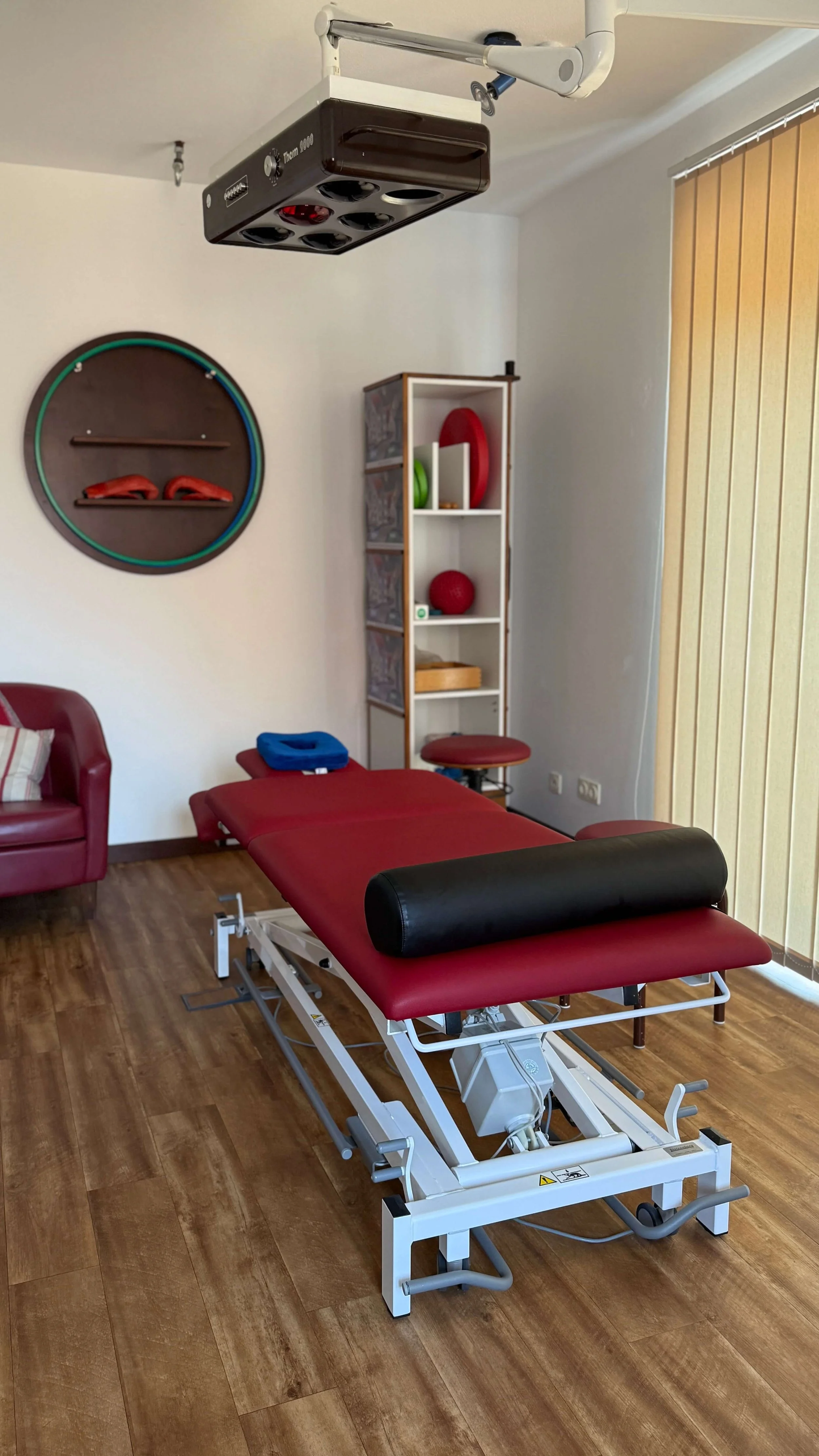 Behandlungsliege in einer Physiotherapiepraxis, an der Wand Hanteln und Bälle zur Rehabilitation, im Hintergrund eine Vitrine mit medizinischen Geräten, Holzboden, vertikaler Sonnenrollo.