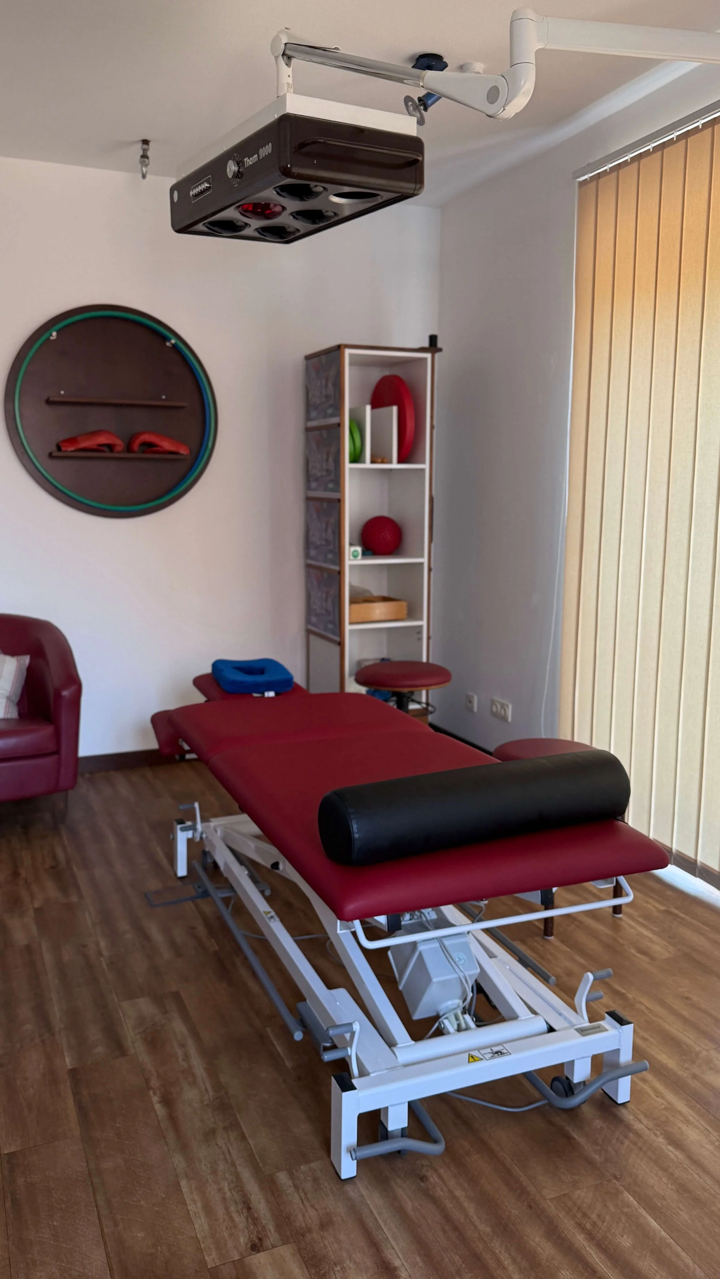 Behandlungsstuhl in einem Therapieraum mit medizinischer Ausrüstung und roten Sitz- und Liegeflächen, heller Raum mit Holzfußboden, Wandregale mit roten und grünen Therapiegeräten, vertikale Jalousien am Fenster.
