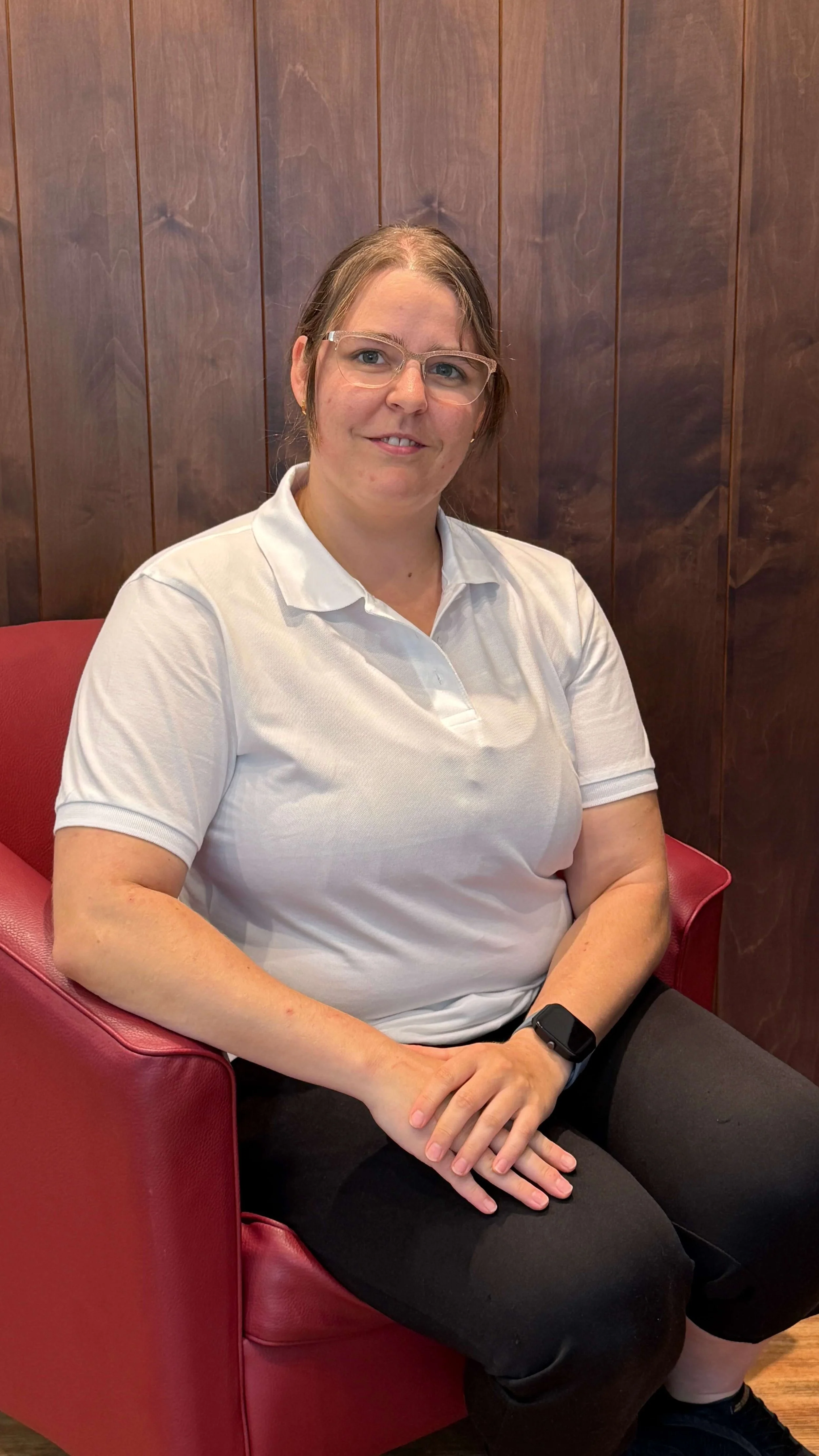 Eine Frau mit Brille und einem weißen Polo sitzt auf einem roten Stuhl vor einer Holzvertäfelung.